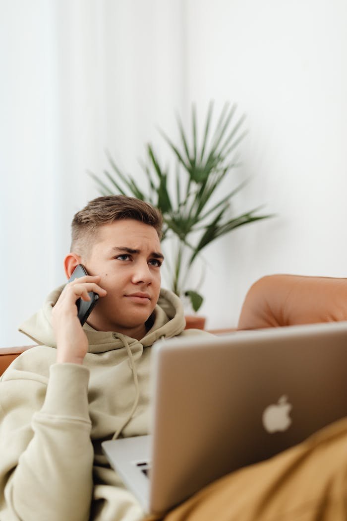 Teen sitting on a couch using a laptop and smartphone in a cozy setting.